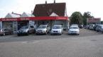 Das Bild zeigt ein Autohaus mit mehreren Autos vor dem Verkaufsbüro und einer Werkstatt mit rotem Vordach.