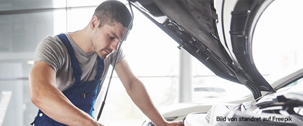 Ein Mechaniker arbeitet am Motorraum eines Autos mit geöffneter Motorhaube in einer Werkstatt.